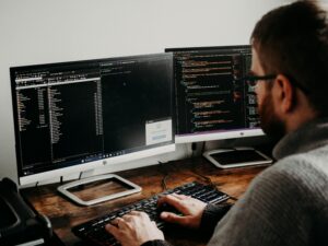 Blonde person with a neck tattoo typing on a keyboard while looking at a monitor displaying computer code in a bright office.