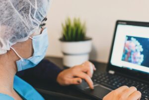Medical professional in a blue scrub top, surgical mask, and hair net using a laptop to research scientific data and microscopic virus images.