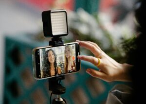 Smartphone on a tripod with an LED ring light filming two women for a video, with a hand reaching to touch the screen interface.