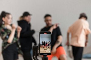 Smartphone mounted on a gimbal stabilizer filming a group of dancers in a studio, with the dance performance visible on the mobile device screen.