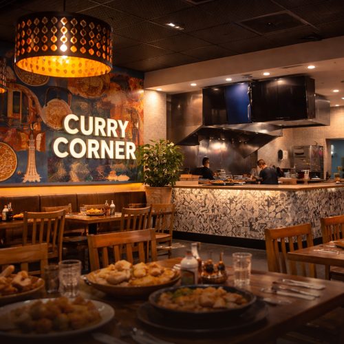 Interior view of "Curry Corner," a modern restaurant with warm lighting, wooden tables set with food, and an open kitchen in the background.