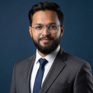 Professional man with a beard and clear-framed glasses wearing a grey suit jacket, white shirt, and navy blue tie against a dark blue background.