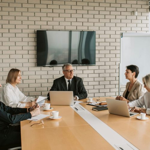 A professional business meeting in a modern office featuring five colleagues in formal attire seated around a light wood conference table with laptops and coffee.